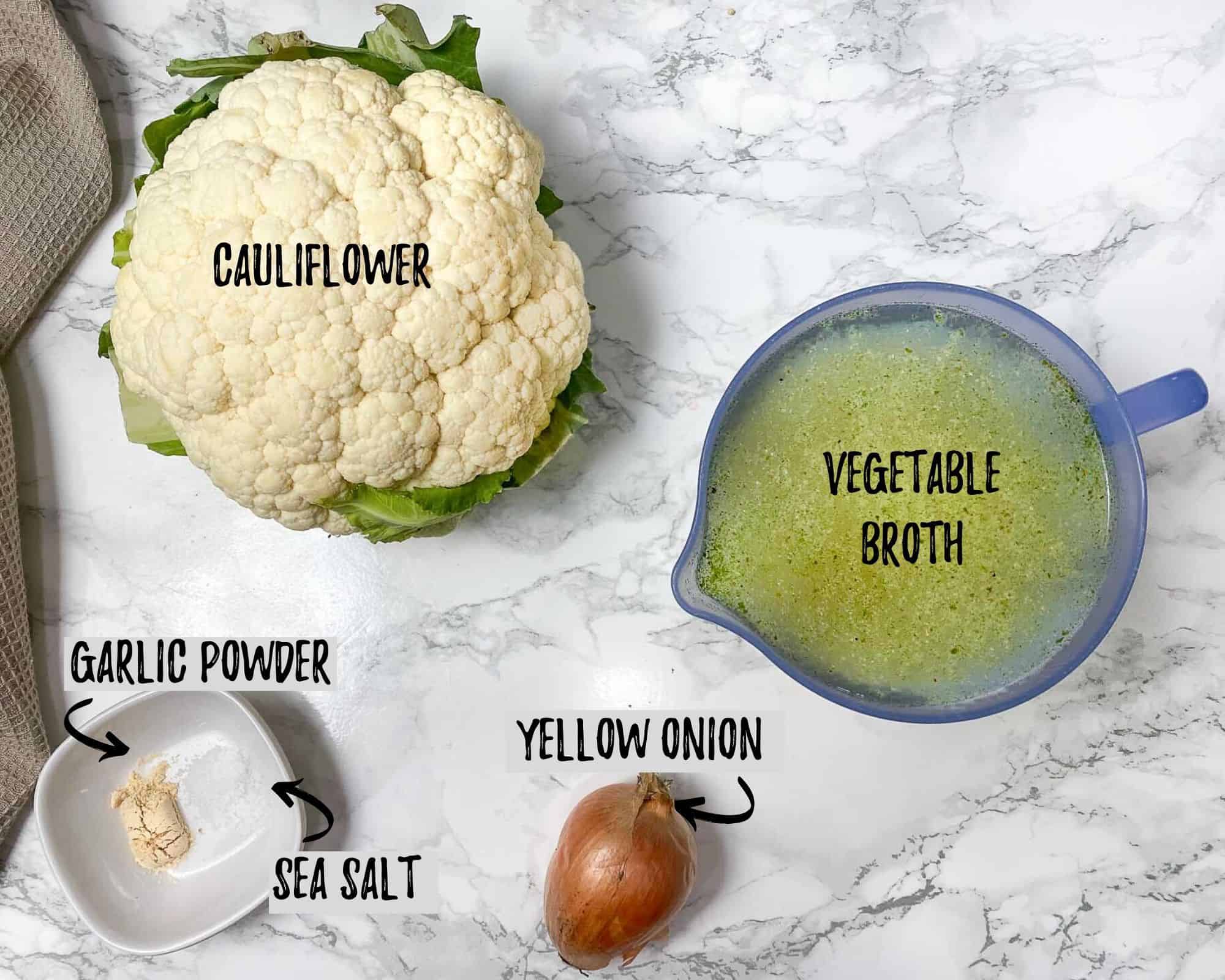 head of cauliflower, yellow onion, bowl of seasoning and measuring cup filled with broth on marble counter