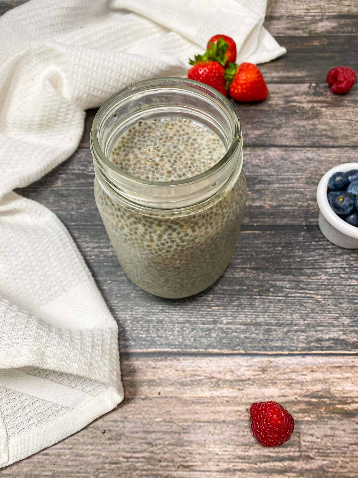 glass jar of chia pudding inside with berries scattered around the jar