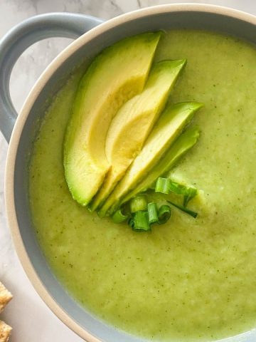 Blue bowl filled with green soup and avocado slices on top.