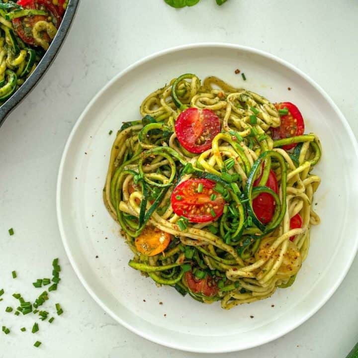White plate with zucchini pasta topped with halved cherry tomatoes.
