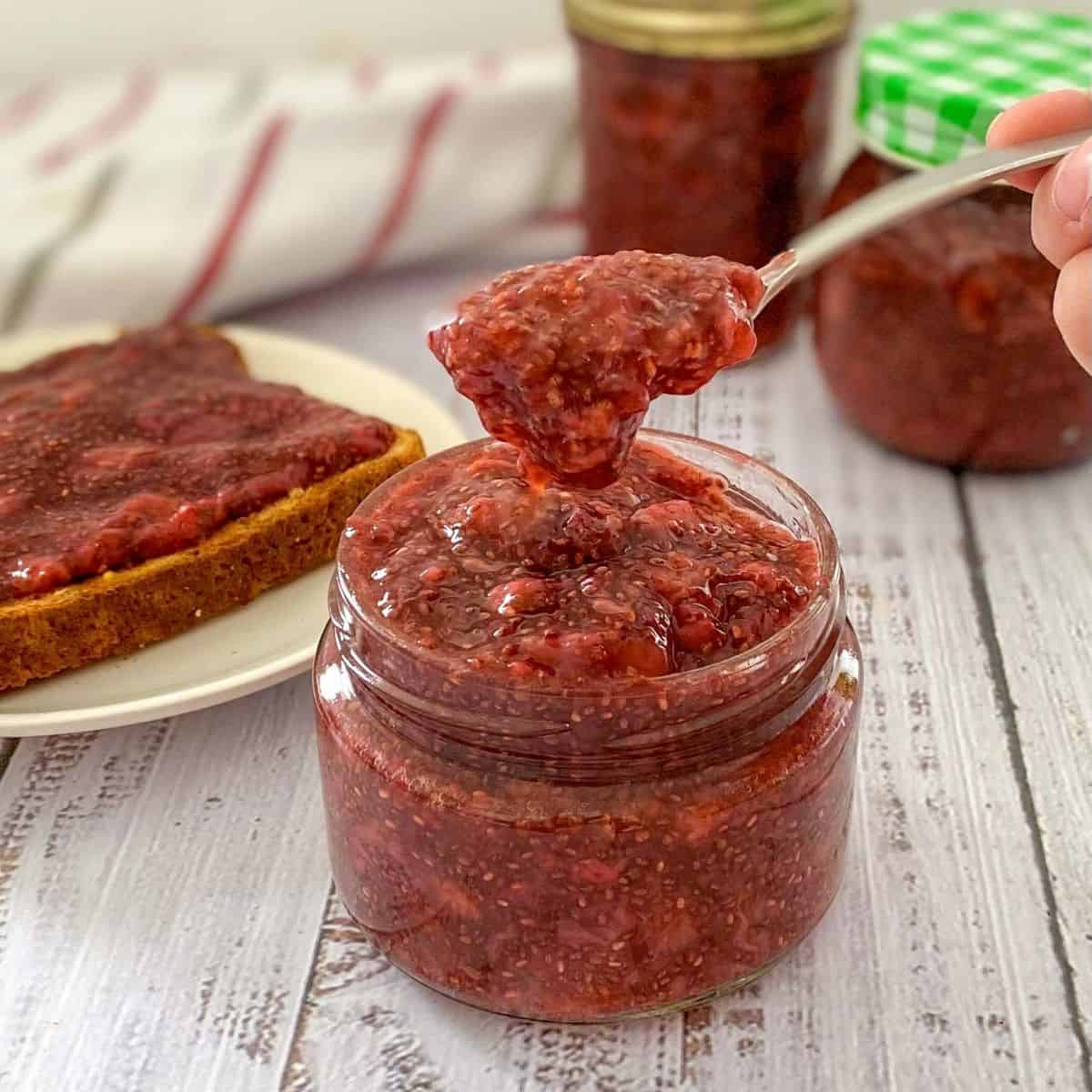 Spoon lifting strawberry jam from glass jar, with more jars of jam in background.