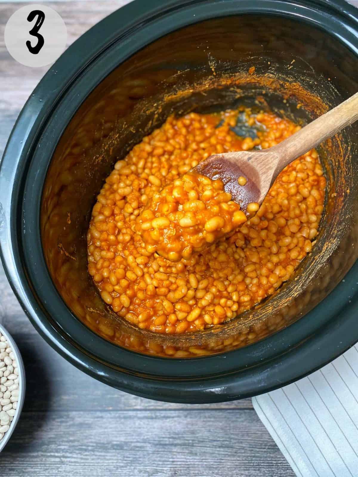 cooked baked beans in slow cooker with wooden spoon lifting up a bite