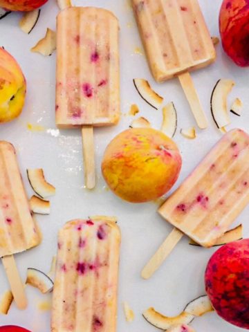 Peach popsicles on white cutting board with coconut smiles and peaches around them.