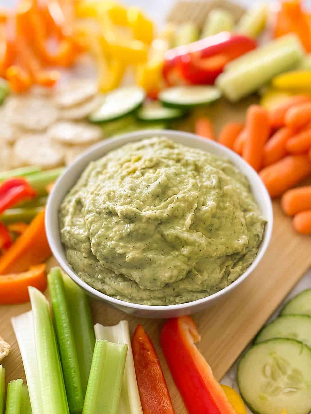 Bowl of dill pickle hummus on cutting board with veggies and crackers around it.
