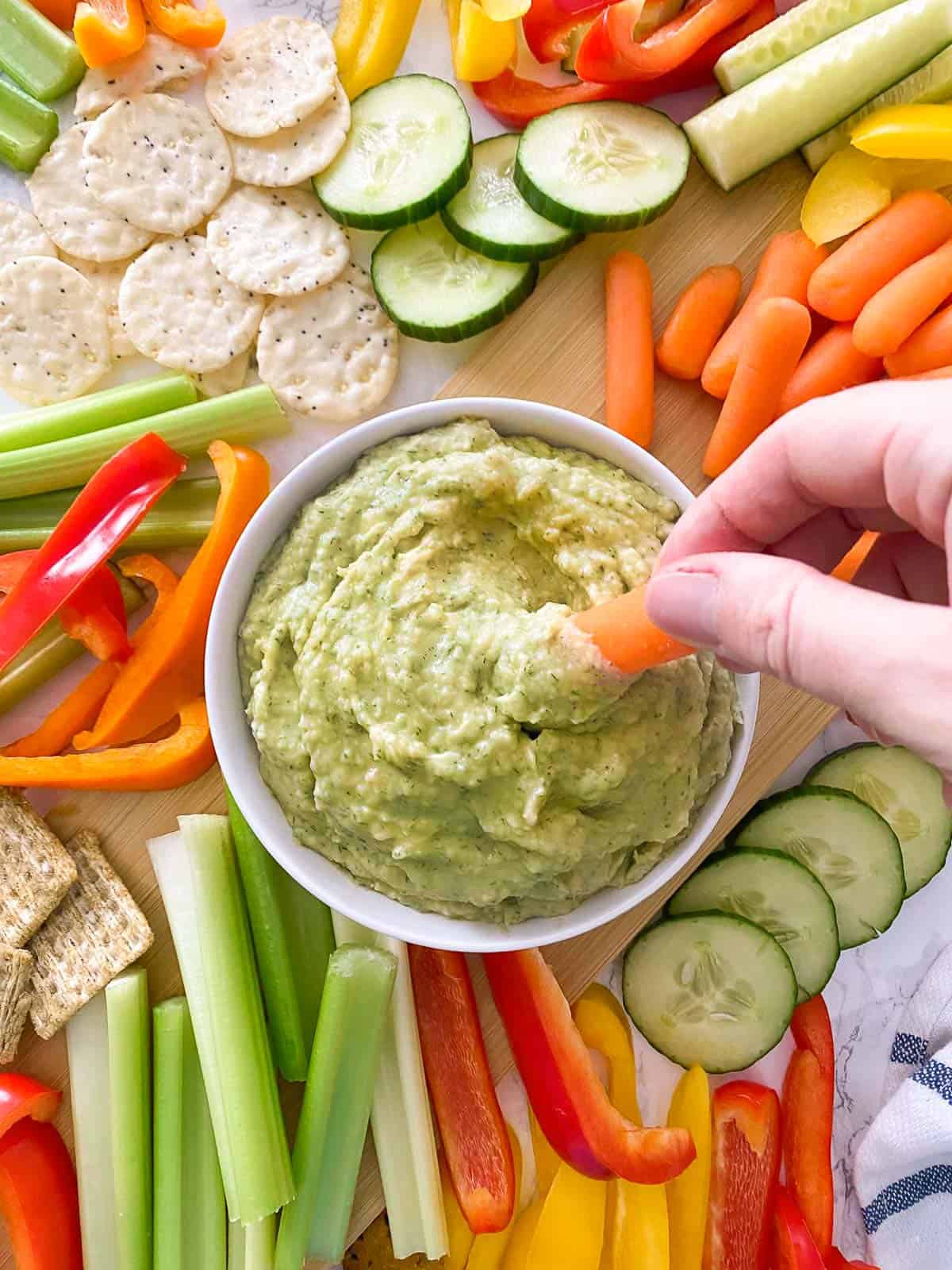 Hand dipping carrot stick into bowl of green dip.