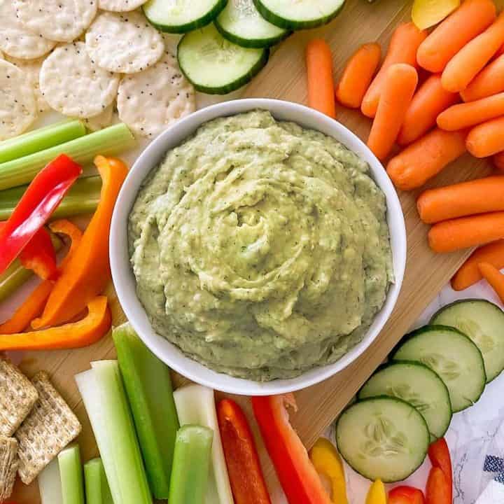 Bowl of dill pickle hummus on cutting board, surrounded by raw veggies.