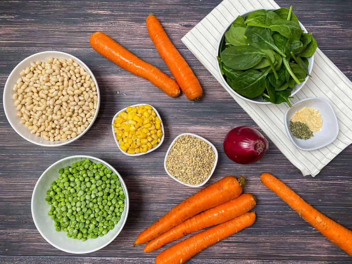grey deck with carrots, red onion, bowl of spinach, peas, barley, beans, and corn scattered around