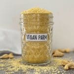 Vegan parmesan cheese overflowing in glass jar with cashews scattered around it.