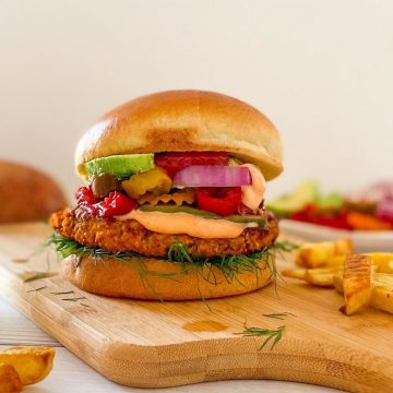 Veggie burger in bun with pickled jalapenos, avocado, onion and ketchup on cutting board.