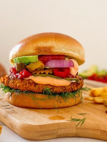 Veggie burger in bun with pickled jalapenos, avocado, onion and ketchup on cutting board.