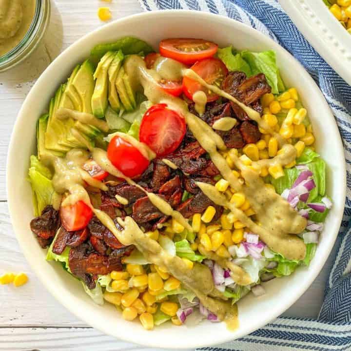 White bowl of salad with rows of avocado, grape tomato, mushrooms, corn and red onion with dressing on top.