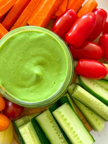 Green dip in glass jar with raw veggies surrounding it.