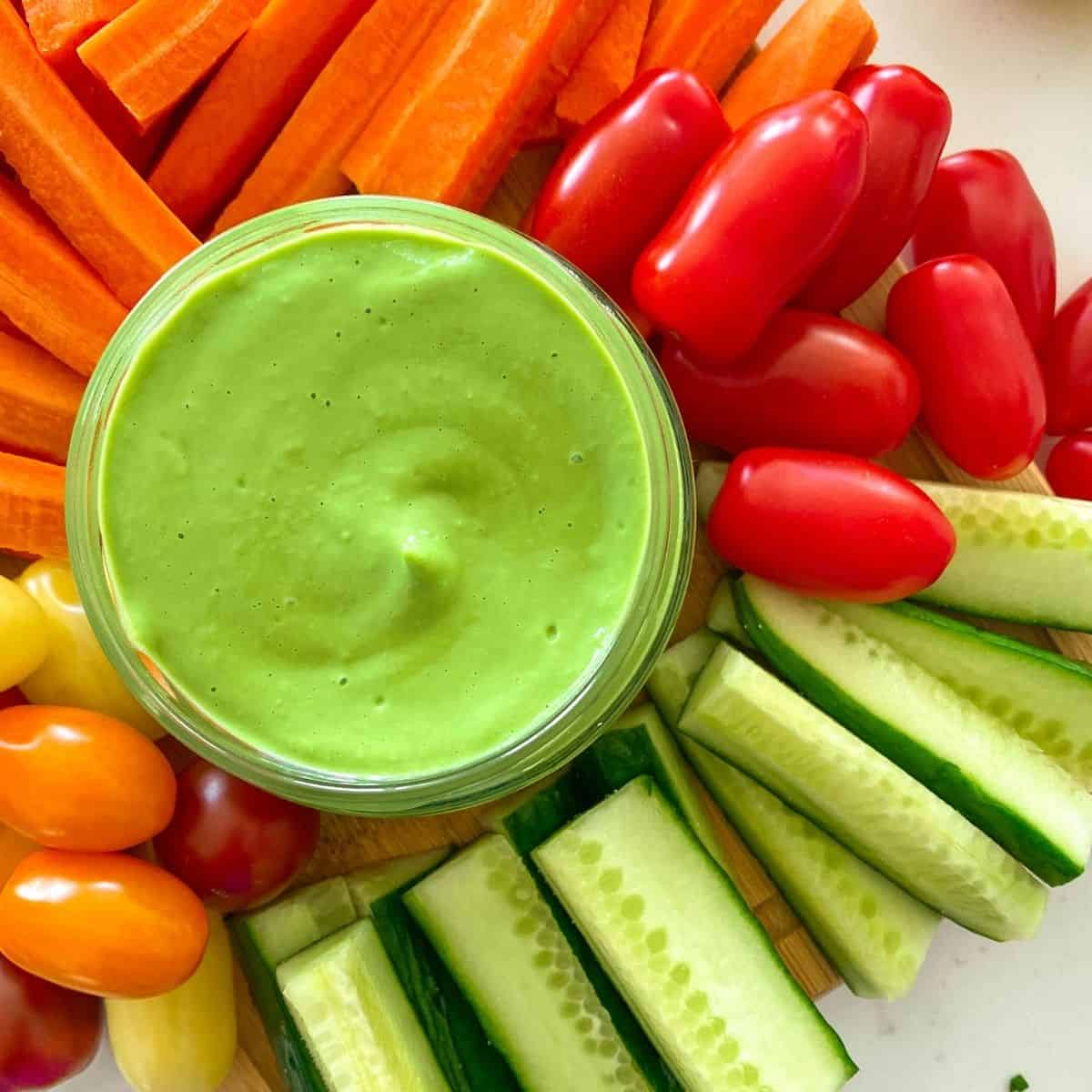 Green dip in glass jar with raw veggies surrounding it.