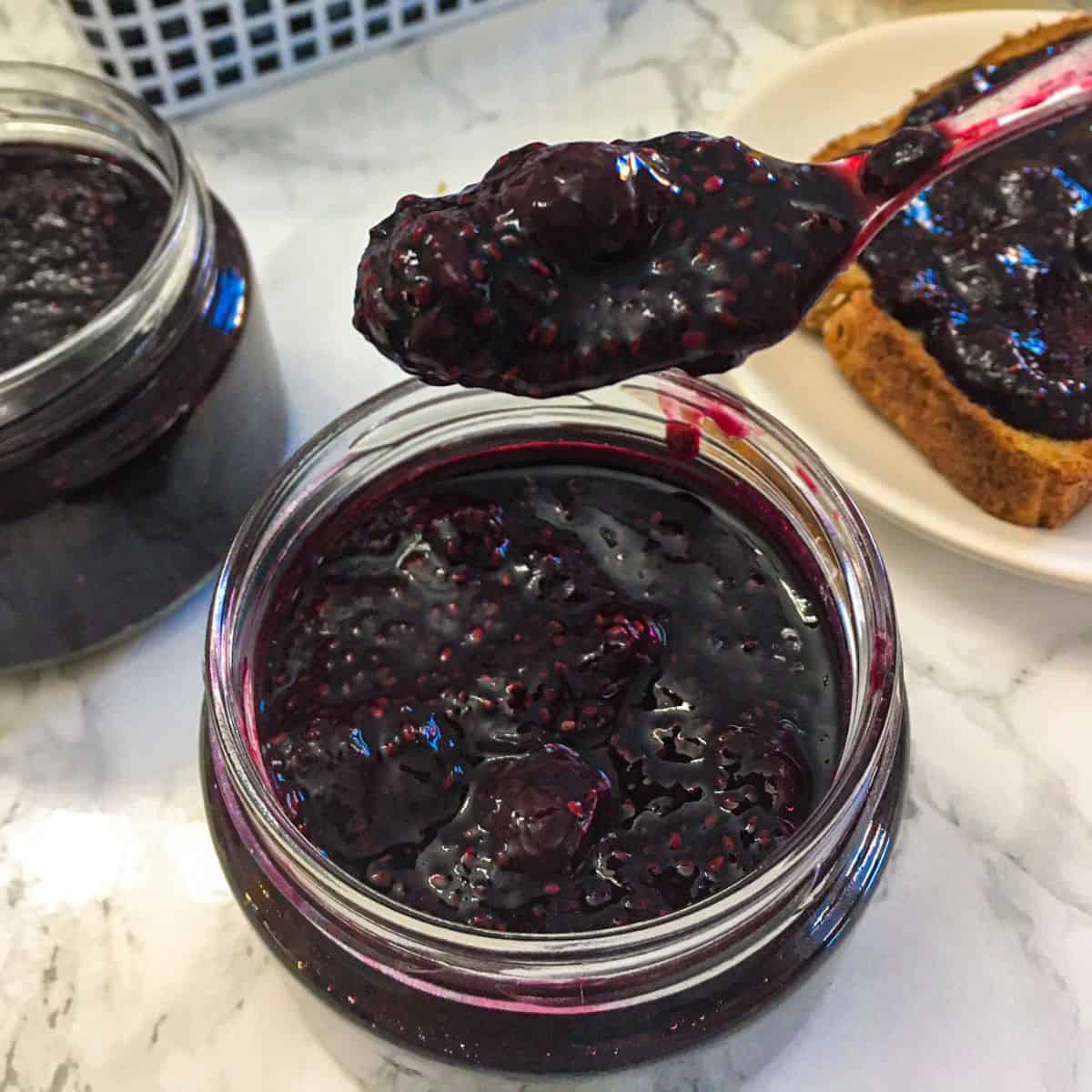 Spoonful of blueberry jam being held above jar of jam.