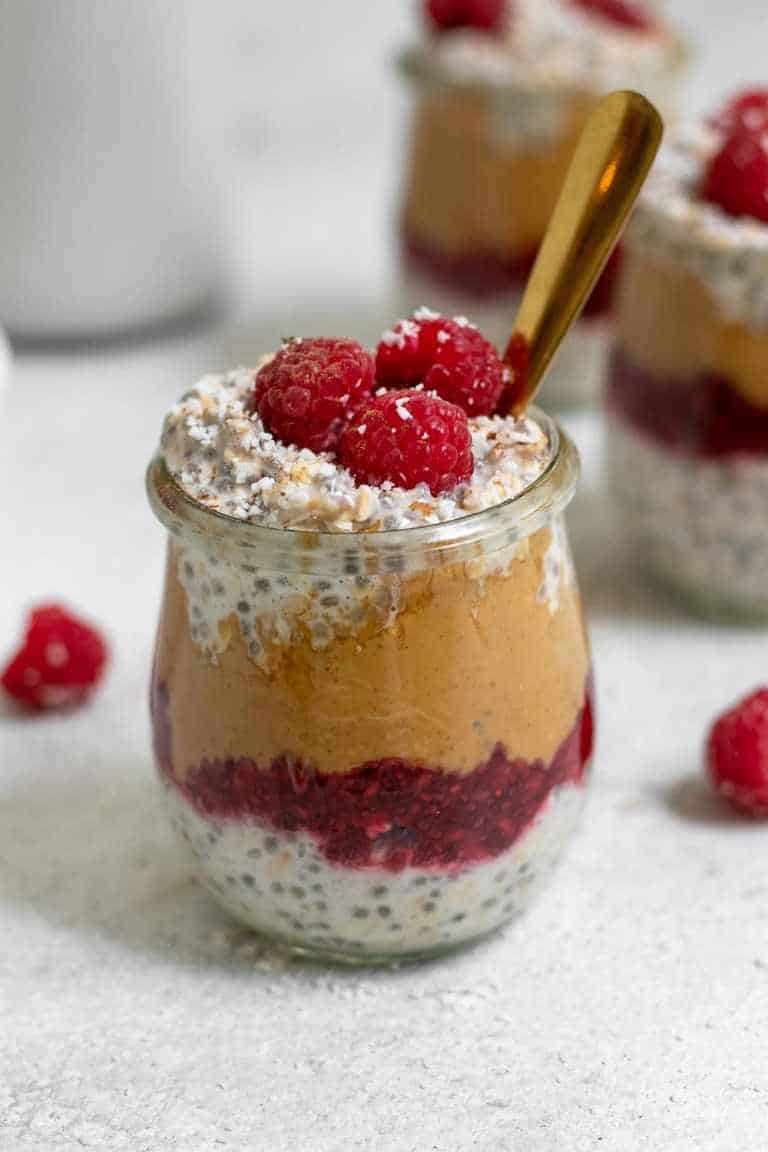 Glass jar of oats with layer of jelly and then peanut butter and raspberries on top.