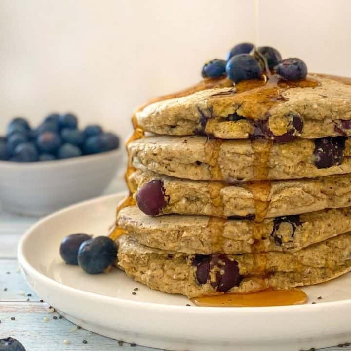 Stack of blueberry pancakes on white plate with blueberries on top and syrup dripping down the sides.