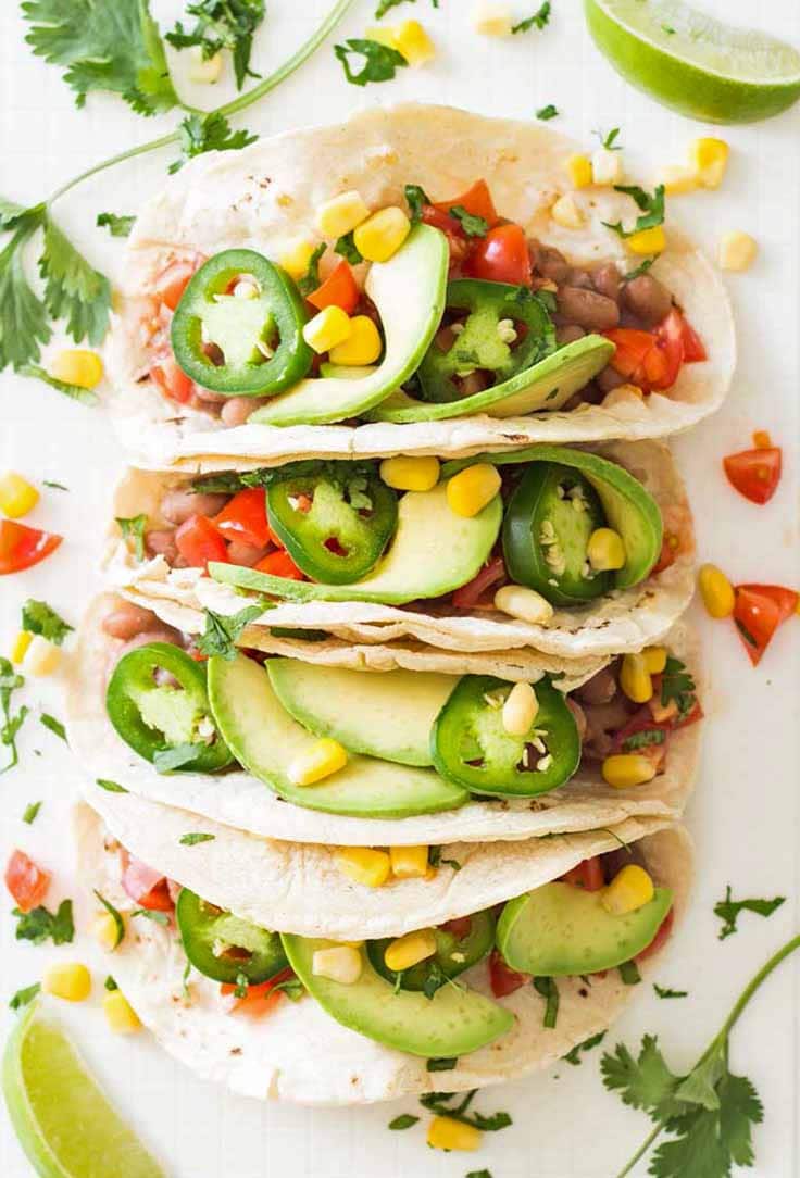 four tacos lined up with pinto beans, avocado, tomato and jalapeno