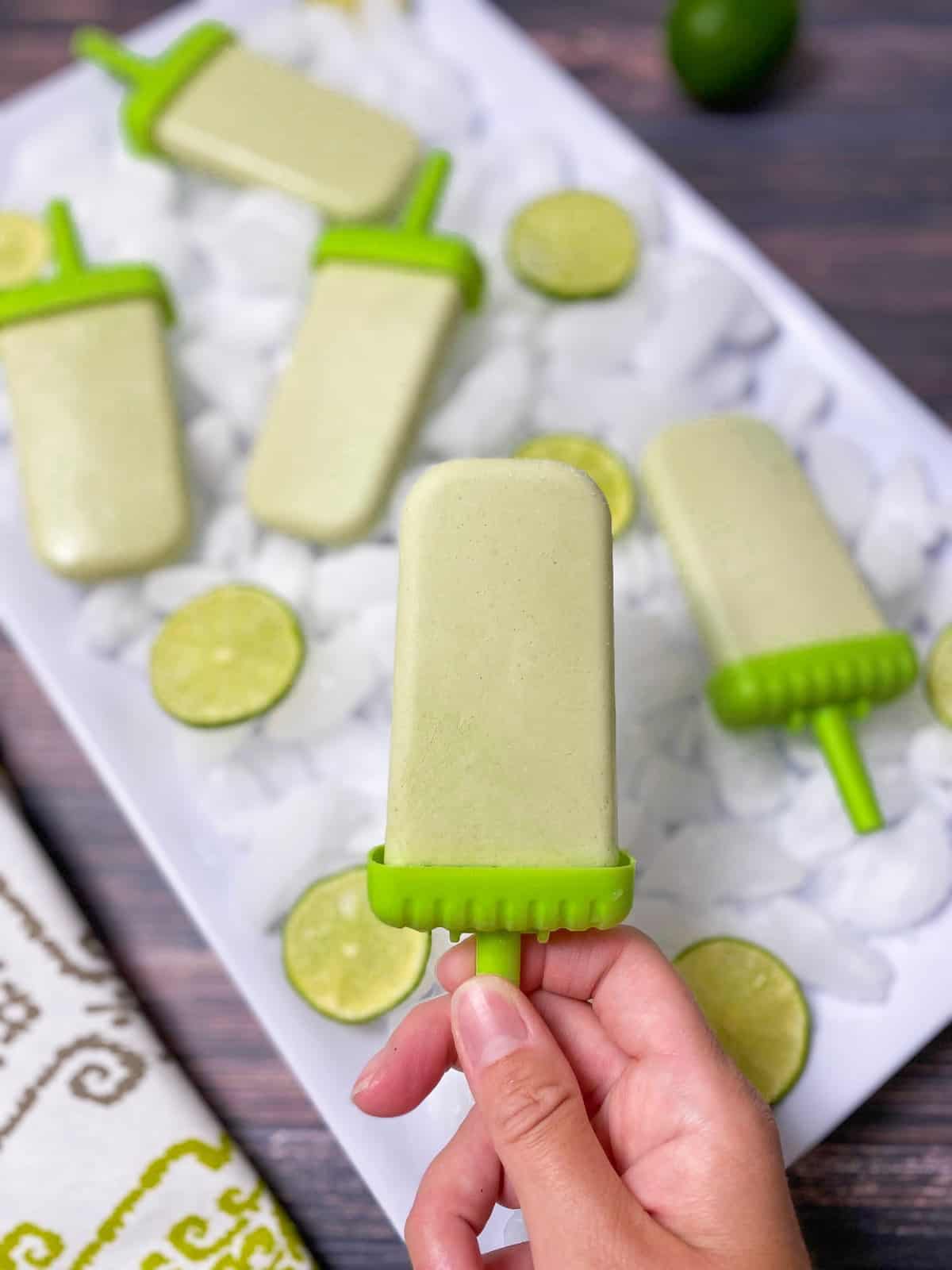 hand holding up lime popsicle with tray of ice and popsicles in the background
