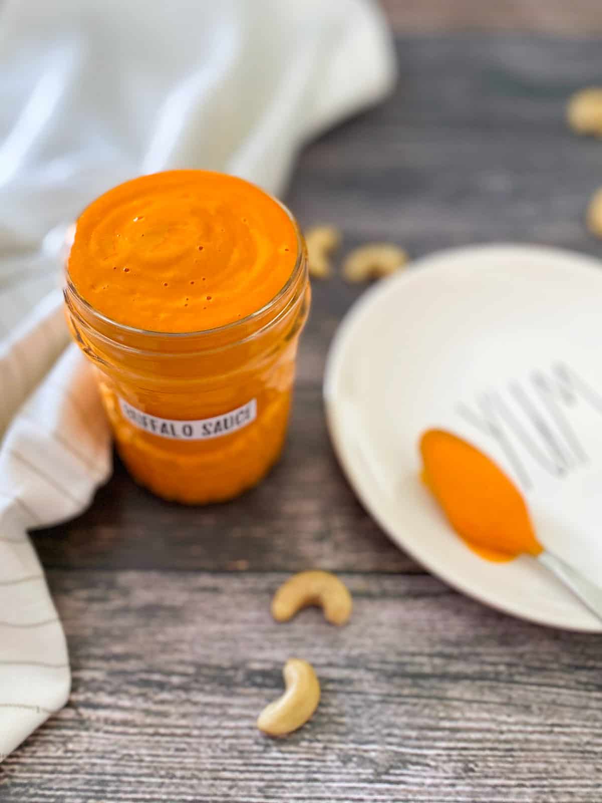 jar of hot sauce with spoonful of sauce on white dish beside the jar and cashews scattered around