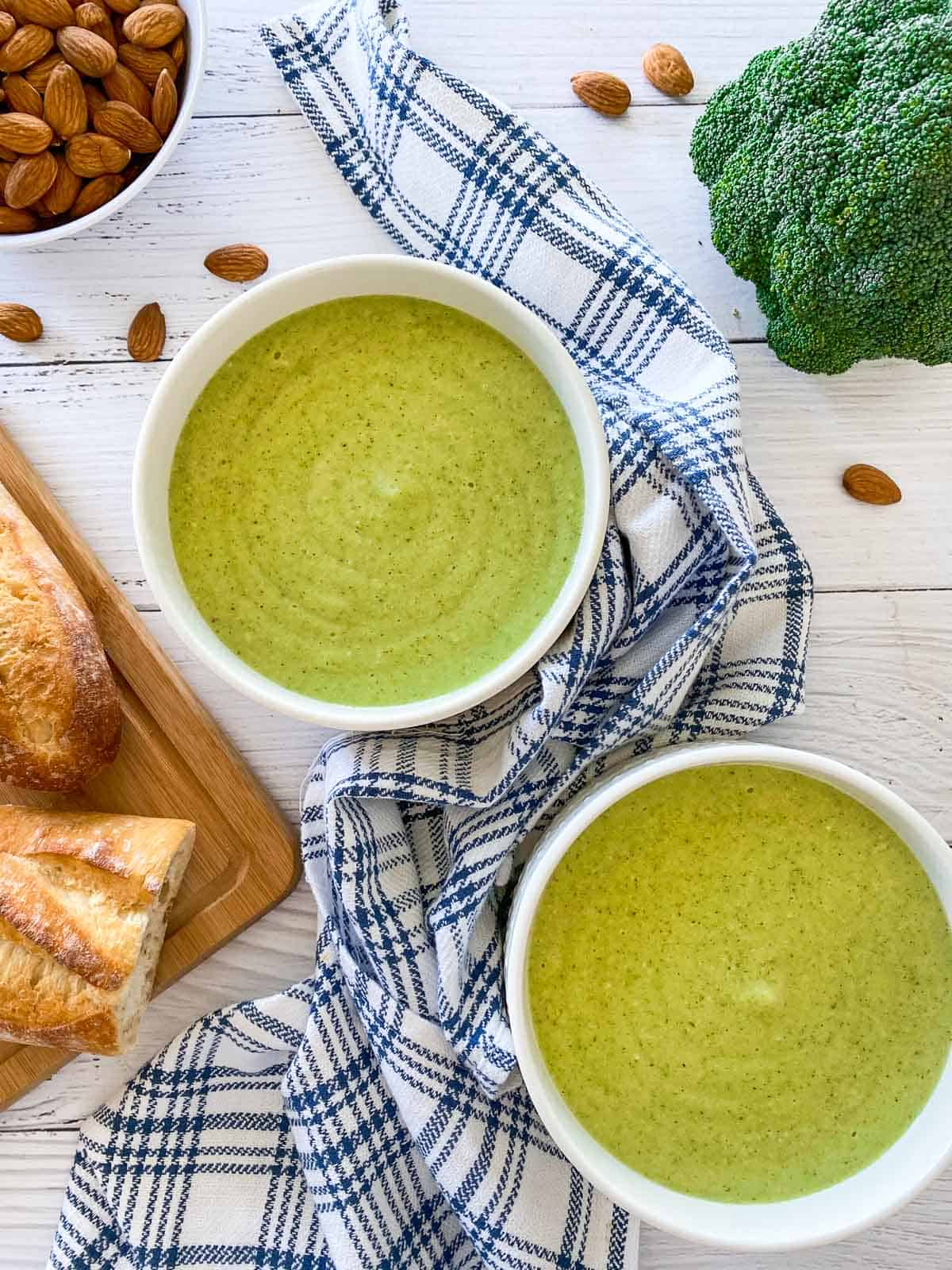 two bowls of green soup with broccoli, almonds and bread in the background.