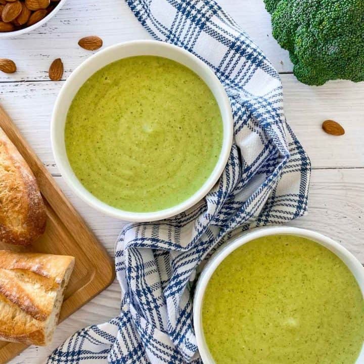 two white bowls of green soup with broccoli and almonds in background.