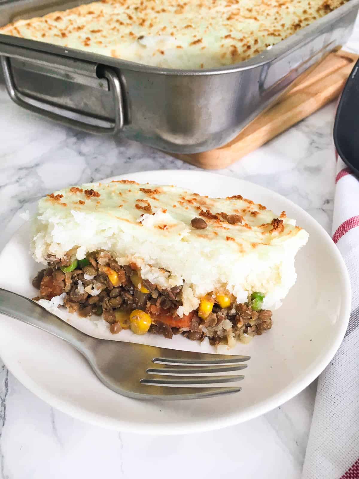 Slice of shepherd's pie in white plate with remaining tray of pie in the background.