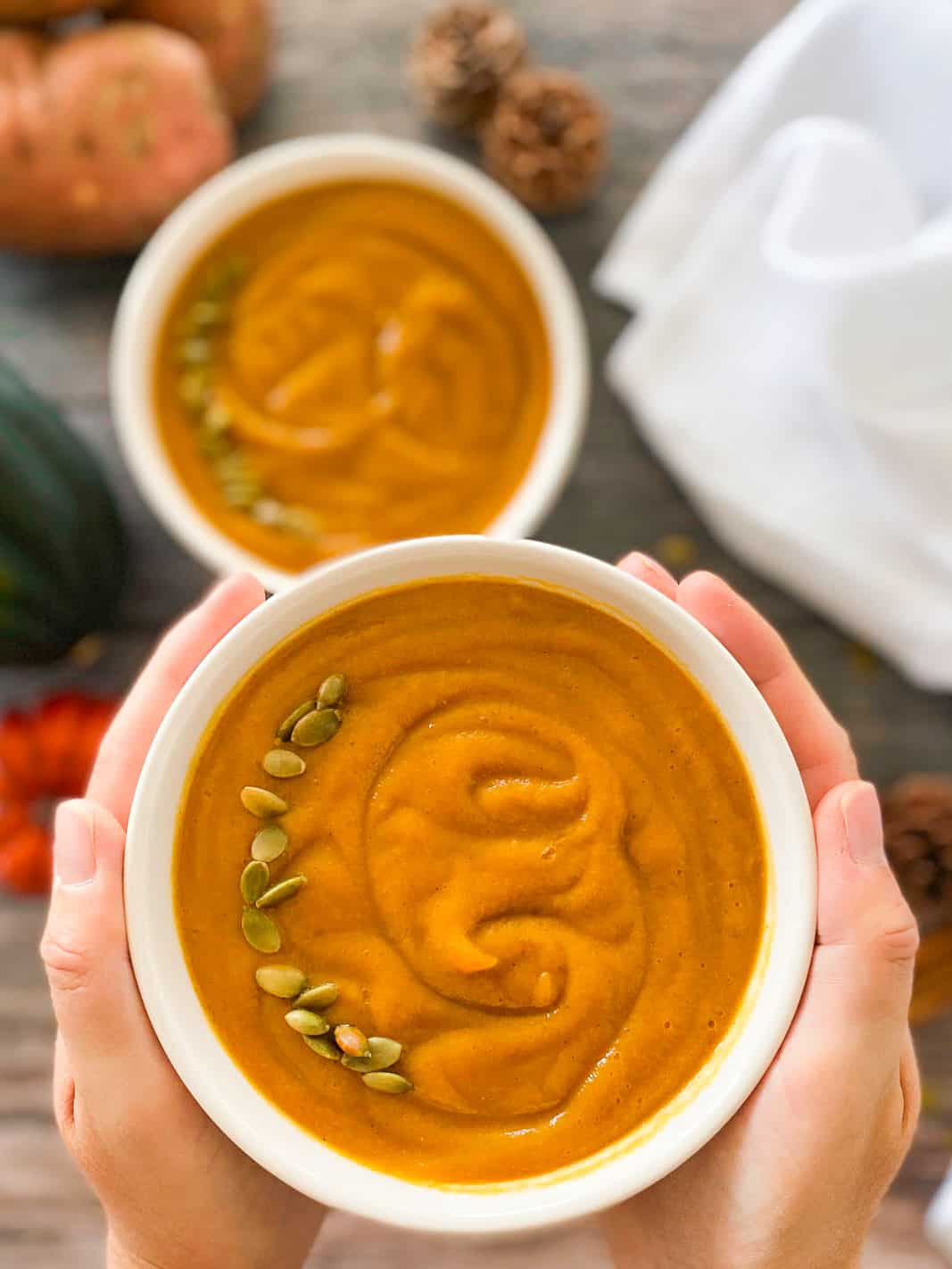 Hand holding up bowl of orange soup with pumpkin seeds on top.