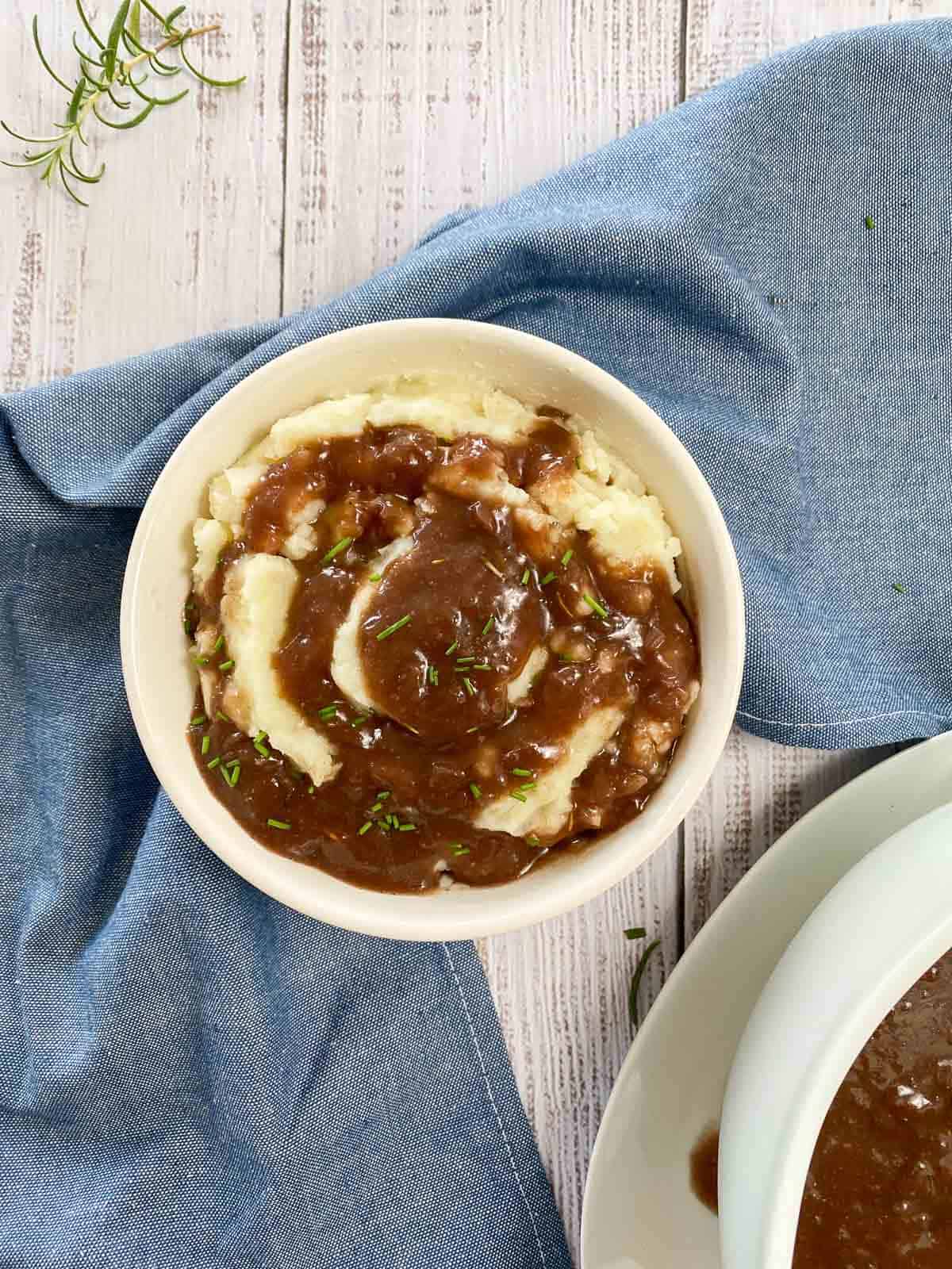 Bowl of mashed potatoes with gravy and chives on top with blue towel underneath.