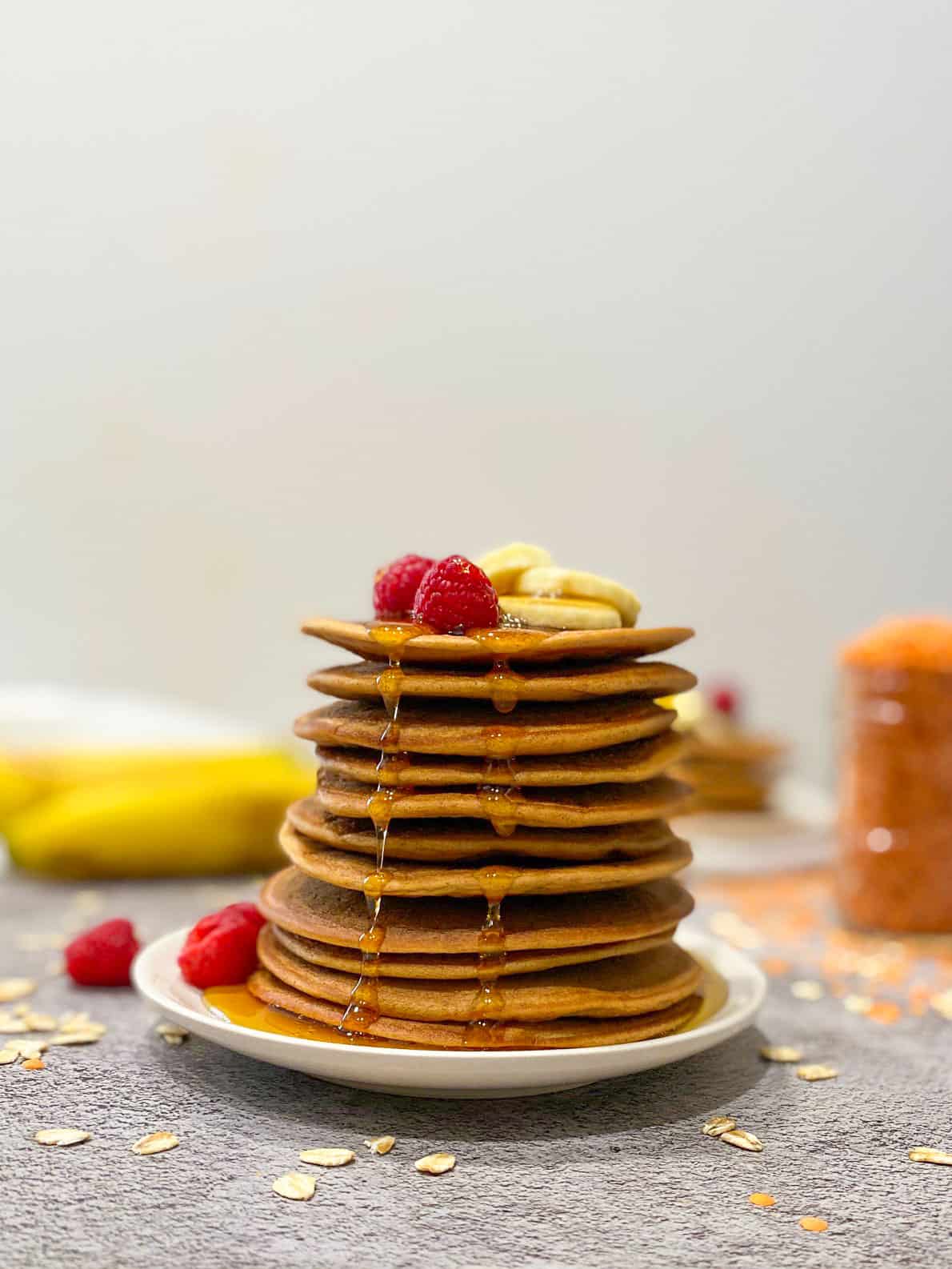 Stack of lentil pancakes with raspberries and banana slices on top and maple syrup dripping down.