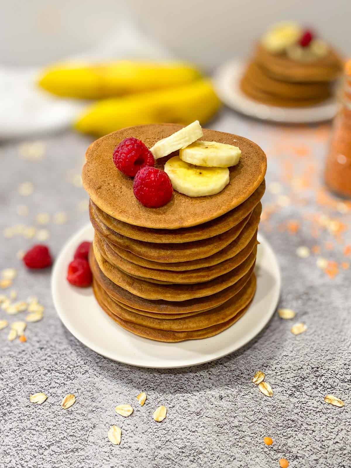 Stack of pancakes on plate with raspberries and banana slices on top.
