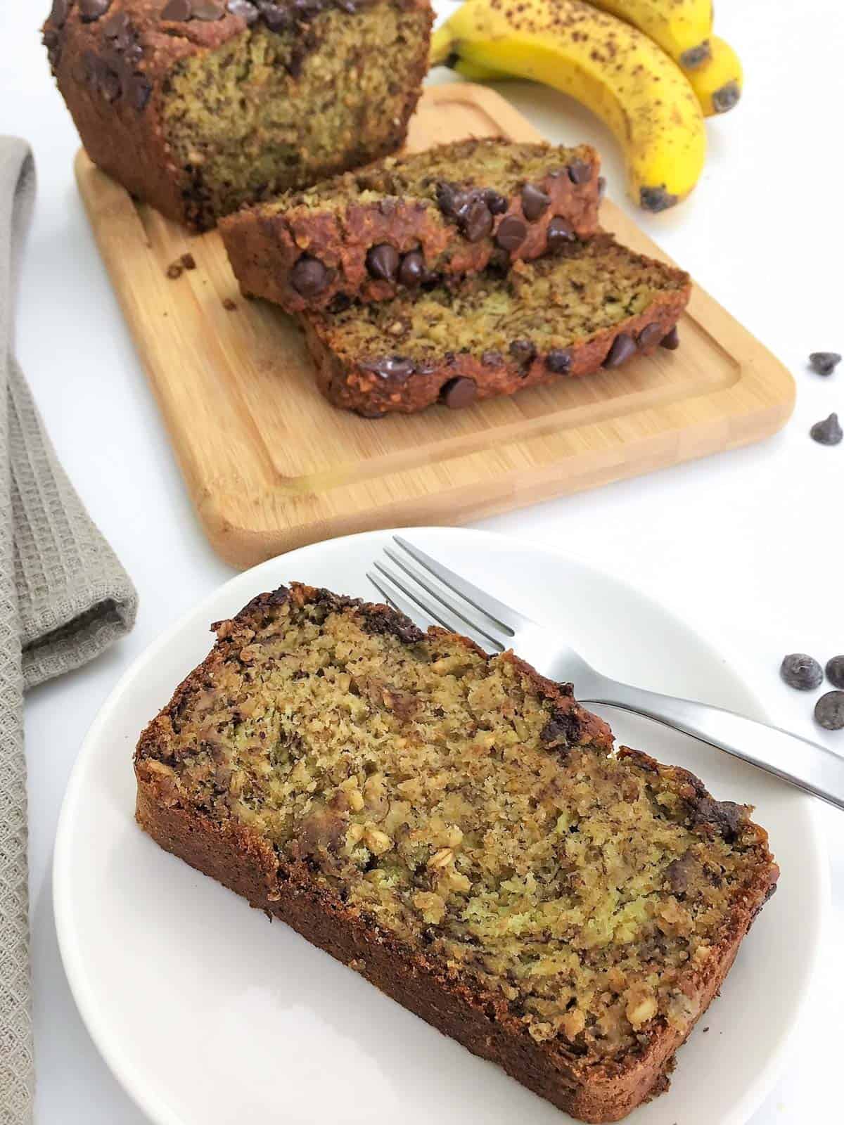 One slice of banana bread on plate with fork and loaf in background.