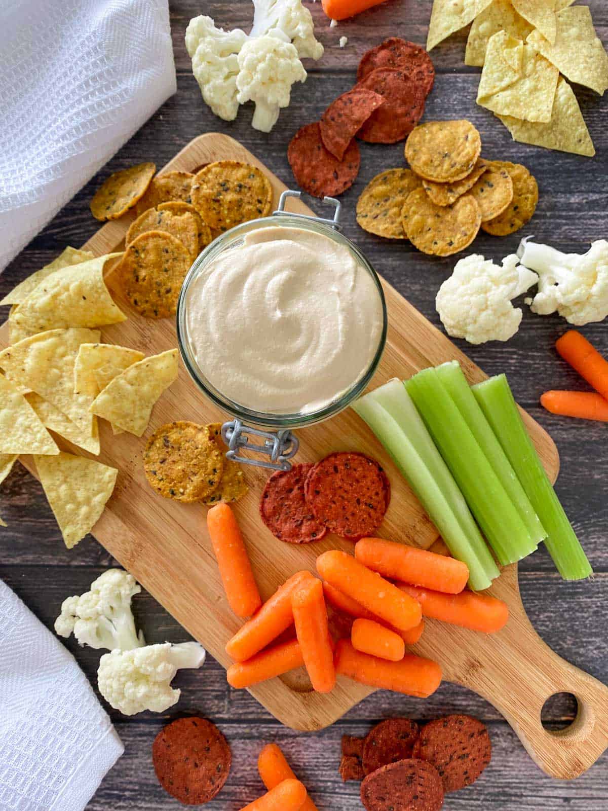 Cutting board with crackers and veggies on top and glass container of vegan cheese dip in the center.