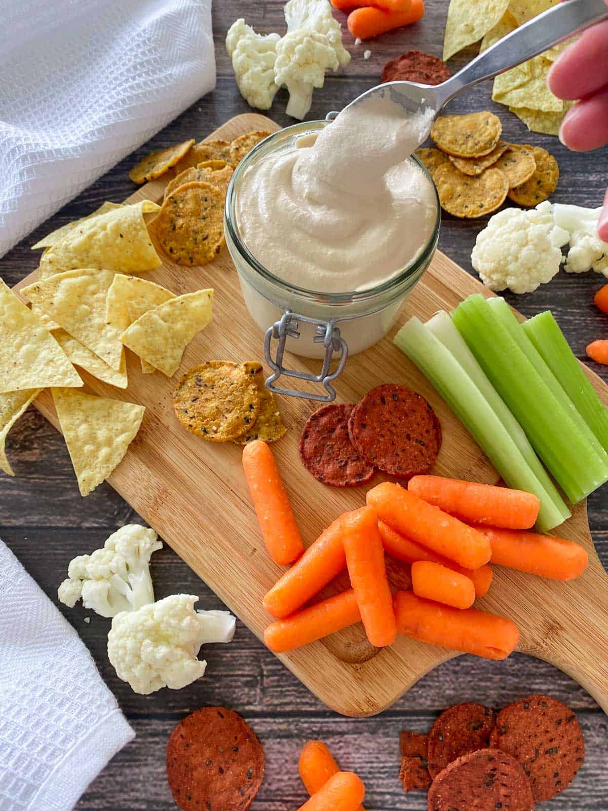 Spoon of walnut cheese sauce dripping into glass jar of more cheese sauce with veggies scattered around the jar.