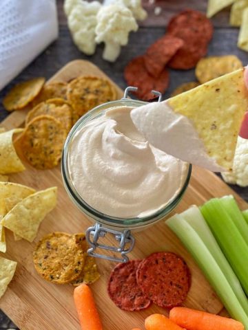 vegan walnut cheese sauce in glass container with hand holding dipped chip above it.