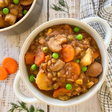 White bowl of stew with mushrooms, carrots, potatoes, peas, lentils, corn.