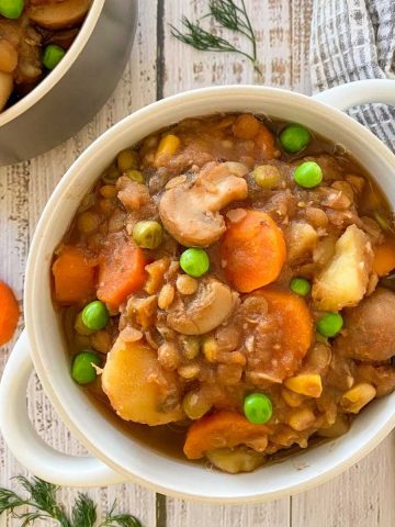 White bowl of stew with mushrooms, carrots, potatoes, peas, lentils, corn.