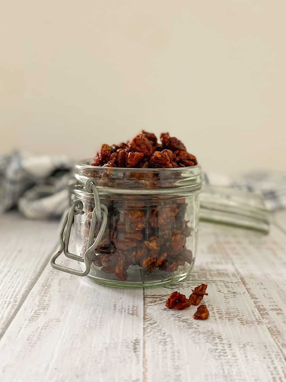 Glass jar filled with candied pecans with blue dishtowel behind it.