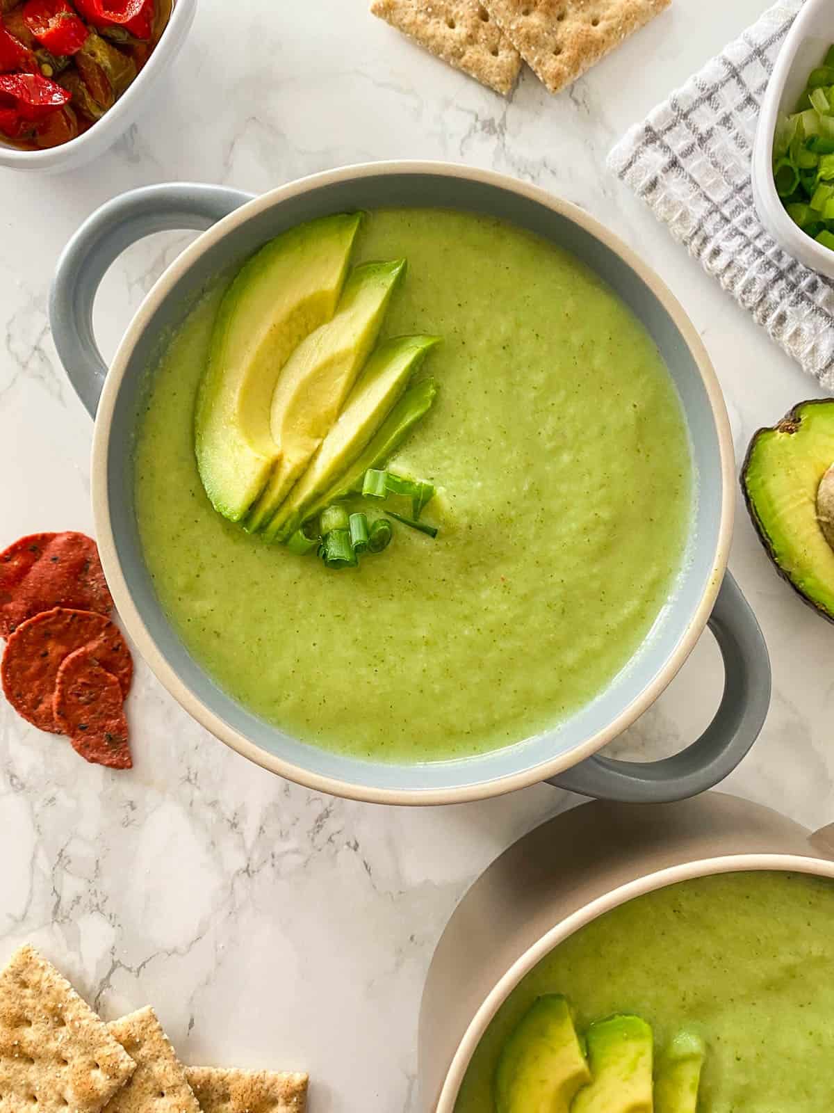 Green creamy soup in bowl with handles and avocado slices on top.