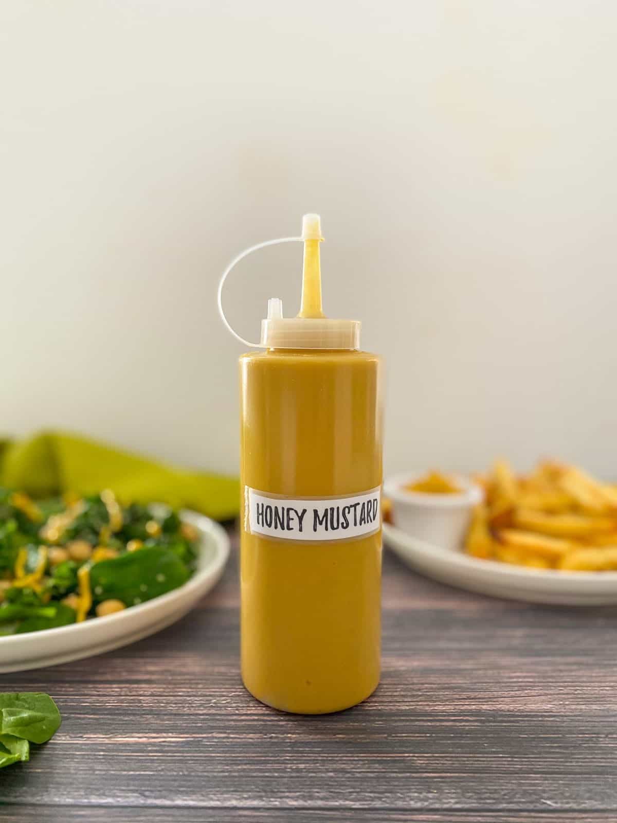 Squeeze bottle filled with yellow dip with plate of salad and fries behind.