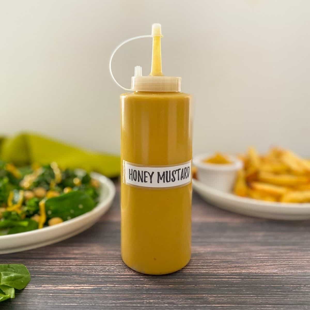 Squeeze bottle filled with yellow dip with plate of salad and fries behind.