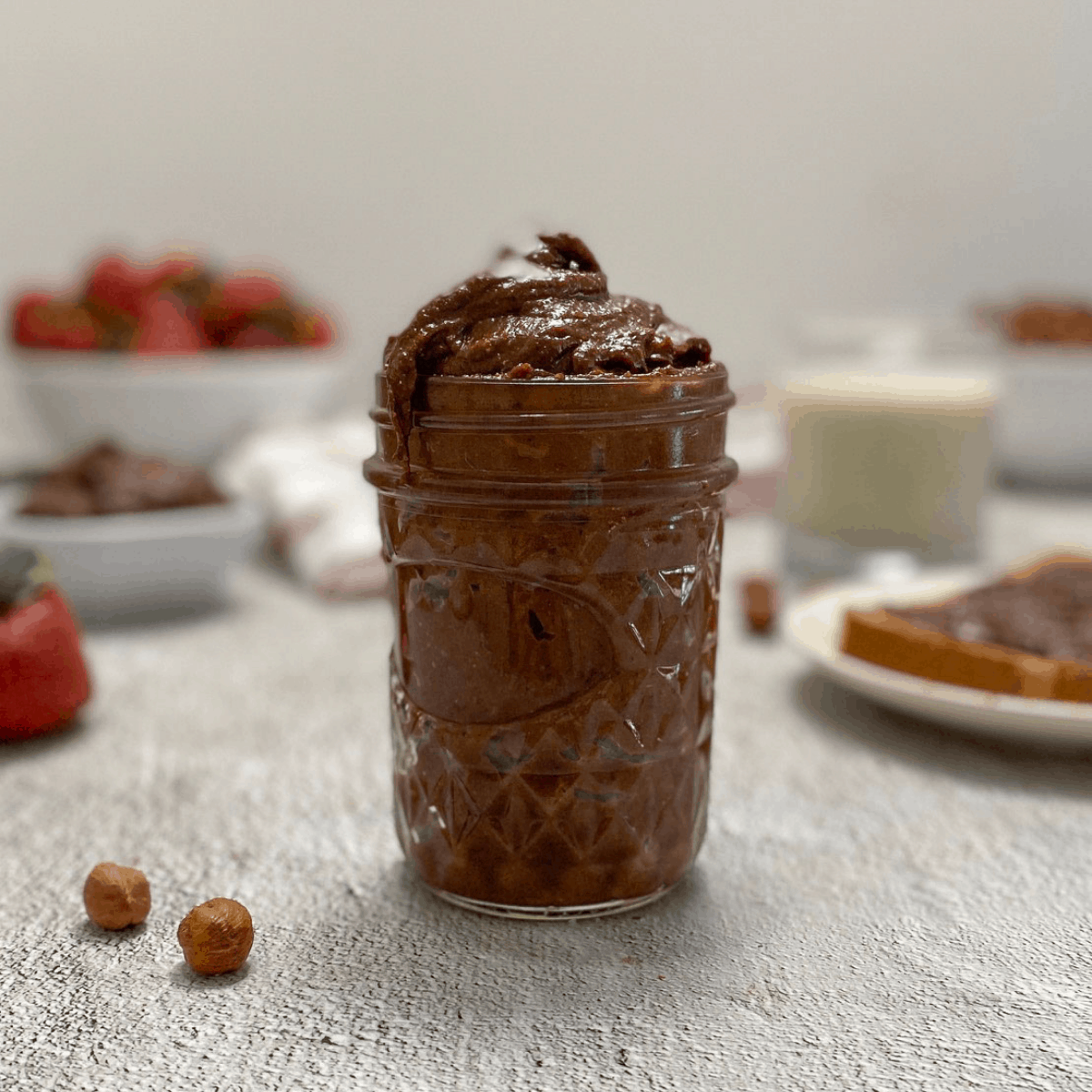 Nutella in glass jar with hazelnuts and strawberries in background.