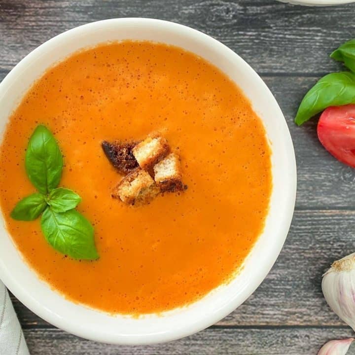 White bowl full of tomato soup with basil and croutons on top.