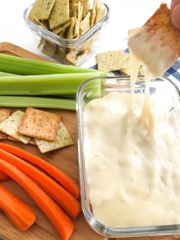 Cracker being dipped into bowl of cheese dip with veggies scattered around it.