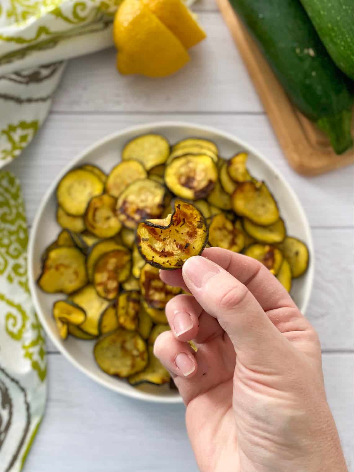 Hand holding up one slice of zucchini with plate of zucchini chips below.