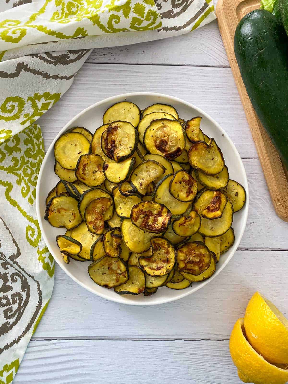 White plate with air fried zucchini chips and zucchini and lemon in the background.