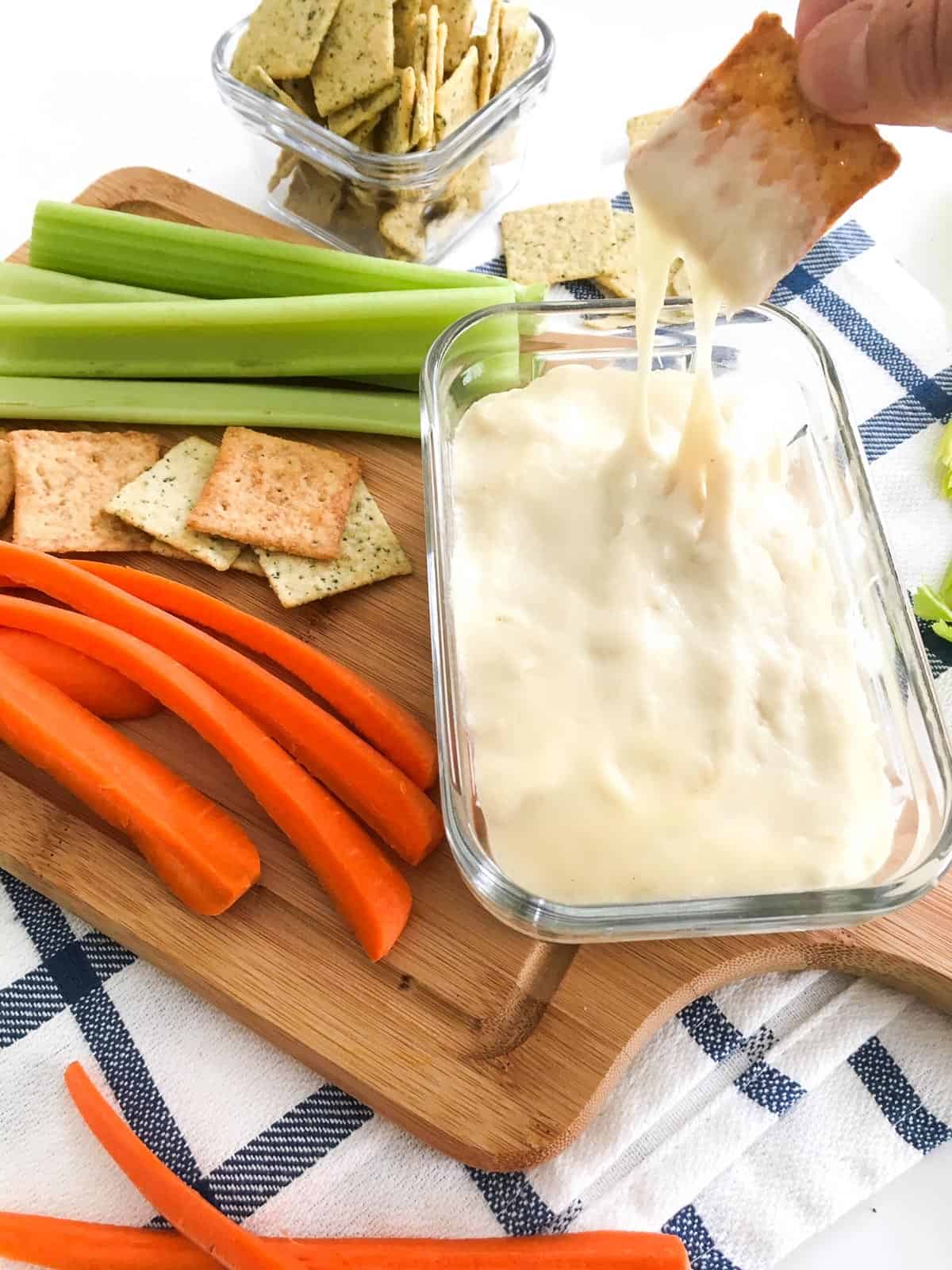 Cracker being dipped into bowl of cheese dip with veggies scattered around it.