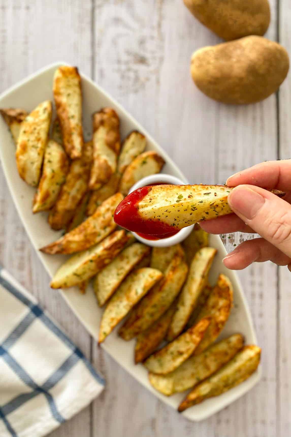 Hand holding up potato wedge dipped in ketchup with more wedges below in platter.