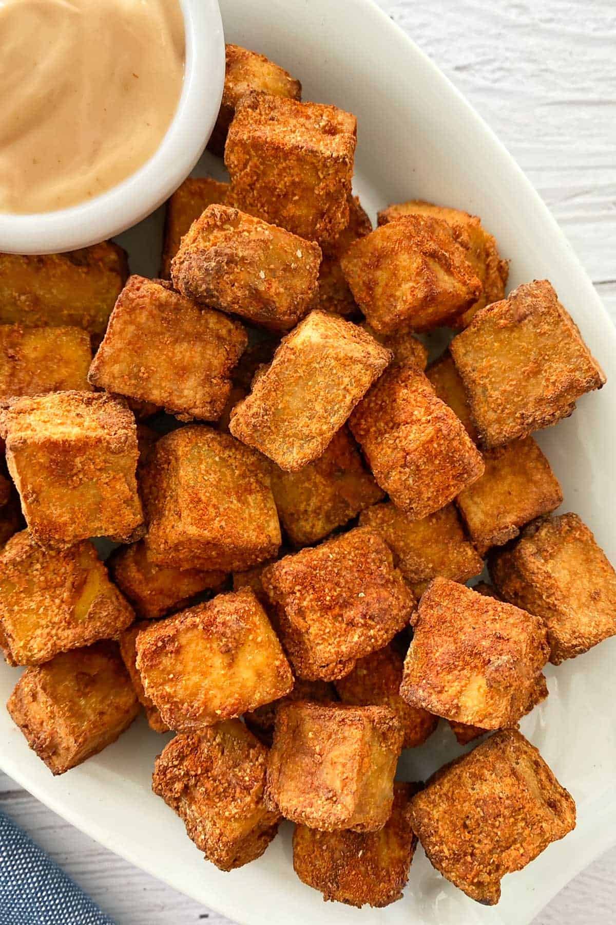 Crispy air fried tofu cubes in white serving dish with dipping sauce on the side.