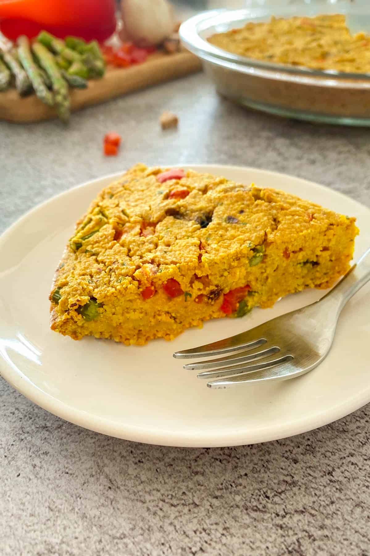 Slice of vegan frittata on small plate with fork, with remaining frittata in background.
