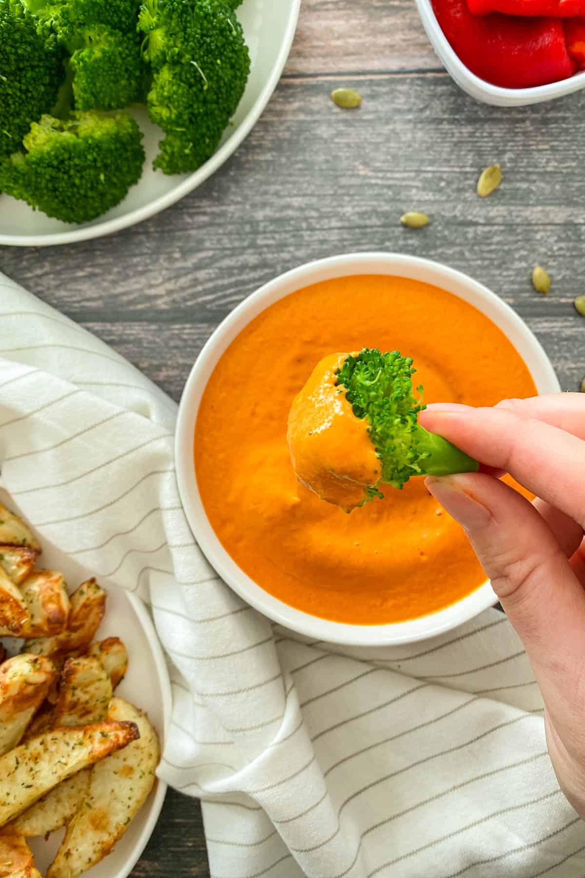 Hand holding broccoli floret dunked in vegan cheese sauce with bowl below.
